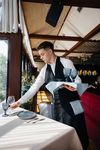 A young male waiter in a stylish uniform is engaged in serving the table in a beautiful gourmet restaurant. Restaurant activity, of the highest level. - Foto, afbeelding