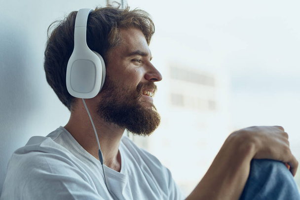 Mann in weißem T-Shirt sitzt mit Kopfhörern am Fenster und hört Musik - Foto, Bild