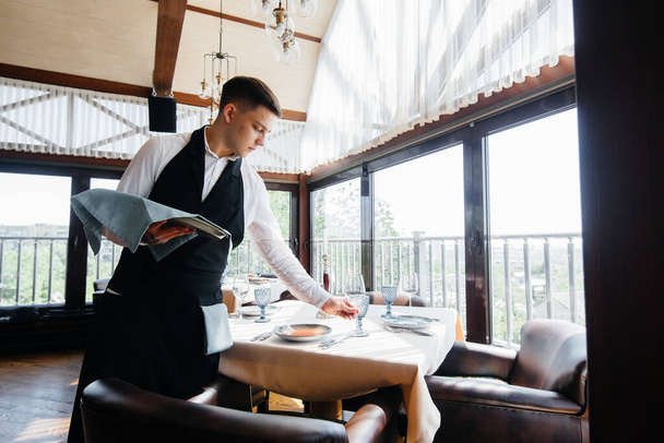 A young male waiter in a stylish uniform is engaged in serving the table in a beautiful gourmet restaurant. Restaurant activity, of the highest level. - Φωτογραφία, εικόνα