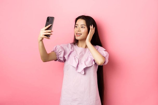 Κομψή Ασιάτισσα blogger που βγάζει selfie στο κινητό, ποζάρει για φωτογραφία smartphone, στέκεται ντυμένη σε ροζ φόντο - Φωτογραφία, εικόνα