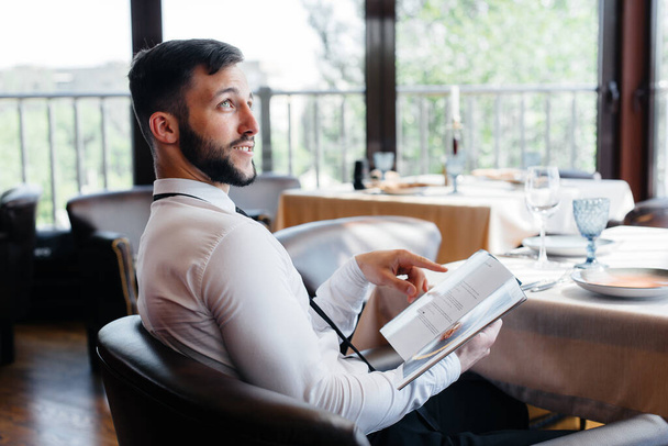 A young businessman in a fine restaurant examines the menu and makes an order to a young waiter in a stylish apron. Customer service. - Foto, Imagen