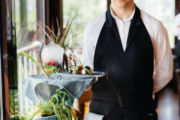 A young waiter in a stylish uniform stands with an exquisite dish on a tray near the table in a beautiful restaurant close-up. Restaurant activity, of the highest level. - Foto, Bild