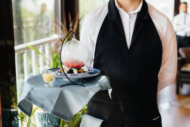 A young waiter in a stylish uniform stands with an exquisite dish on a tray near the table in a beautiful restaurant close-up. Restaurant activity, of the highest level. - Foto, Imagem