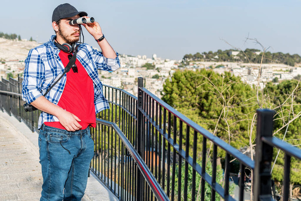 A young attractive bearded Middle Eastern man looking through binoculars while standing on a bridge - Photo, image