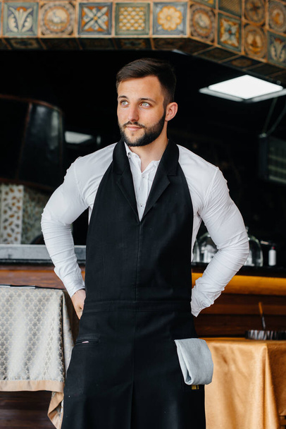 A young waiter with a beard puts on an apron and prepares for a working day in a fine restaurant. - Foto, Bild