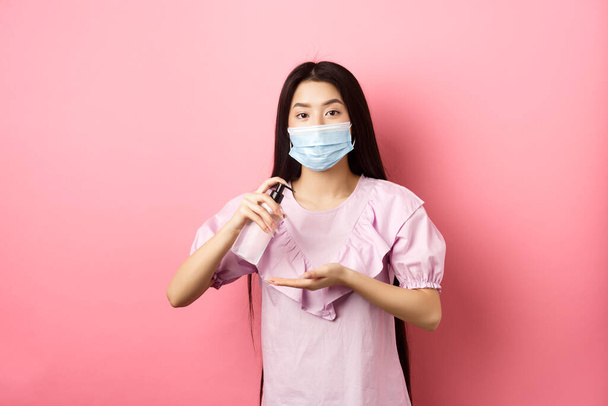 Terveet ihmiset ja covid-19 pandemia käsite. Söpö aasialainen nainen lääketieteellisessä naamiossa puhtaat kädet antiseptisellä, puhdistimella, seisoo vaaleanpunaista taustaa vasten - Valokuva, kuva