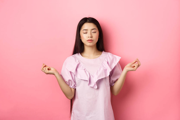 Stay calm. Peaceful asian woman meditating, hold hans in zen gseture and close eyes, practice yoga, standing patient on pink background - Photo, Image