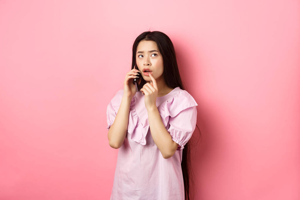 Przemyślana azjatycka kobieta rozmawiająca przez telefon komórkowy i myśląca, dotykająca ust i patrząca w górę zamyślona, stojąca na różowym tle - Zdjęcie, obraz