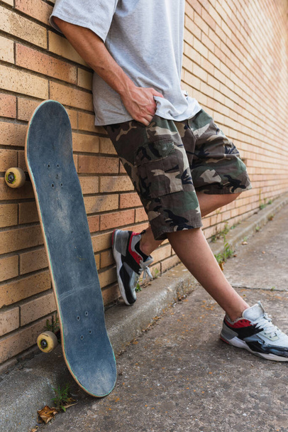 A vertical shot of a young male with his skateboard leaning against a brick wall - Фото, изображение
