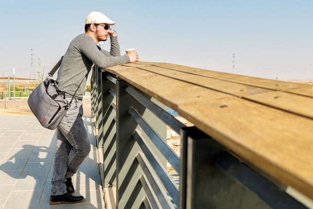 A side view of relaxed man, drinking coffee, waiting on a bridge, on a sunny spring day. - Fotó, kép