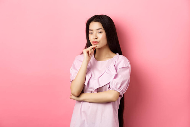 Hmm, interesting. Pensive and cunning asian girl having idea, plan to do something, looking at camera and touching chin with thoughtful face, standing on pink background - Photo, Image