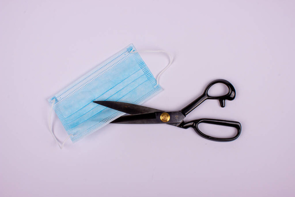A top view of  metal scissors with a protective facemask on a white surface - Foto, imagen