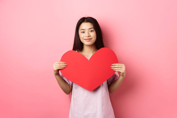Valentines day concept. Cute teenage asian girl showing big red heart card, falling in love, going on romantic date in dress, smiling tender at camera, pink background - Foto, afbeelding