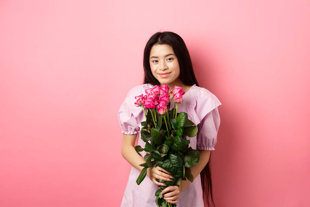 Romantikus érzékeny ázsiai lány gazdaság csokor rózsa, mosolygós aranyos a kamera, miután Valentin-nap szeretője, ruha viselése, rózsaszín háttér - Fotó, kép