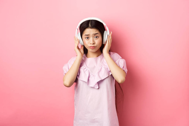 Σκεπτικό κορίτσι χωρίς διασκέδαση με το τραγούδι, ακούγοντας μουσική στα ακουστικά και συνοφρυωμένη δυσαρέσκεια, στέκεται απέναντι σε ροζ φόντο - Φωτογραφία, εικόνα