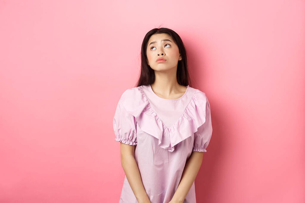 Мечтательная и грустная азиатка смотрит на логотип верхнего левого угла, стоя задумчивый и мрачный в розовом романтическом платье, белый фон - Фото, изображение