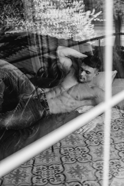 A young sexy athlete with perfect abs is lying on the bed in the studio topless in jeans. Healthy lifestyle, proper nutrition, training programs and nutrition for weight loss. Black and white. - Fotografie, Obrázek