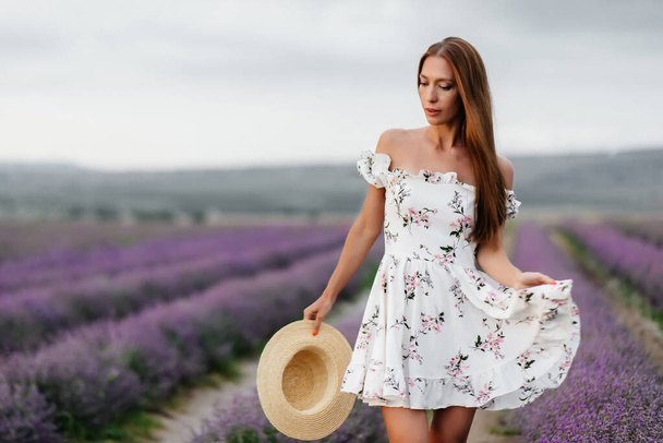 A young beautiful girl in a delicate dress and hat walks through a beautiful field of lavender and enjoys the aroma of flowers. Vacation and beautiful nature. - Foto, Imagem