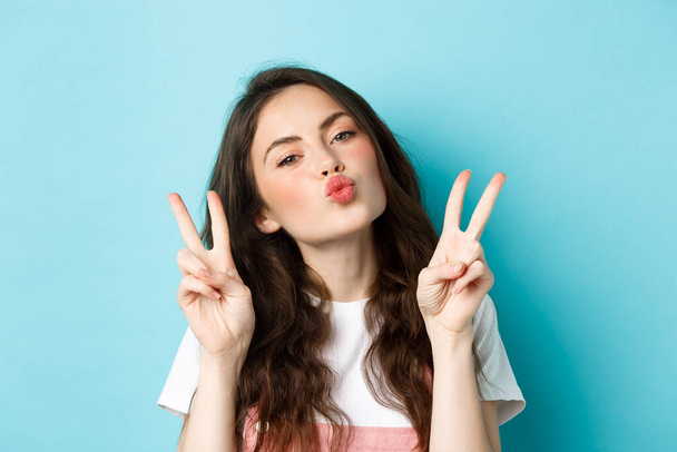 Close up portrait of glamour brunette girl making kissing face, showing kawaii v-sign and smiling cute at camera, standing happy against blue background - Фото, изображение