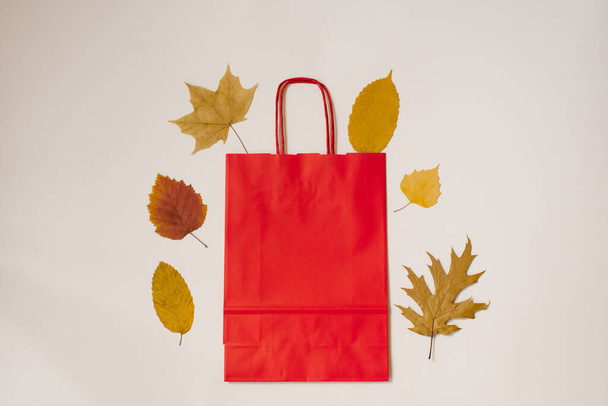 秋のフラットレイアウト。薄い背景に工芸品の赤い袋と秋の落ち葉が並んでいます。割引・販売の秋のコンセプト - 写真・画像