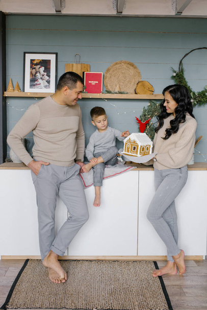 Eine stylische junge Familie mit kleinem Kind steht in ihrer weihnachtlich dekorierten Küche und bewundert ein selbst gebackenes Lebkuchenhaus. - Foto, Bild