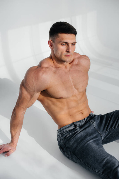 A young sexy athlete with perfect abs sits on the floor in the studio topless in jeans in the background. Healthy lifestyle, proper nutrition, training programs and nutrition for weight loss. - Foto, Imagen