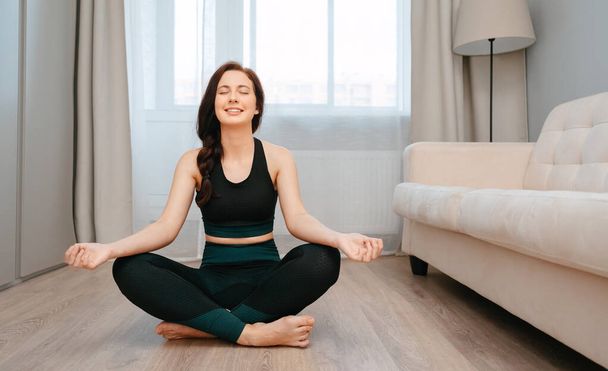 Smiling woman is sitting in the lotus position doing stretching at home - Φωτογραφία, εικόνα