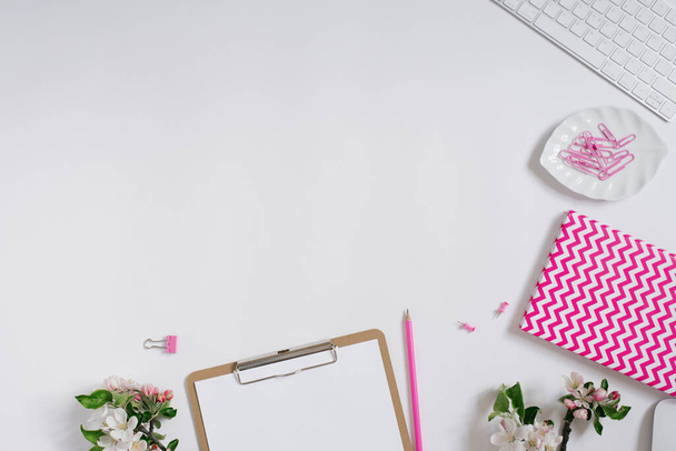 Thuiskantoor bureau werkplek met blanco papier klembord, notitieblok, toetsenbord, briefpapier en appelboom bloemen op witte achtergrond met kopieerruimte. Platte indeling, bovenaanzicht - Foto, afbeelding
