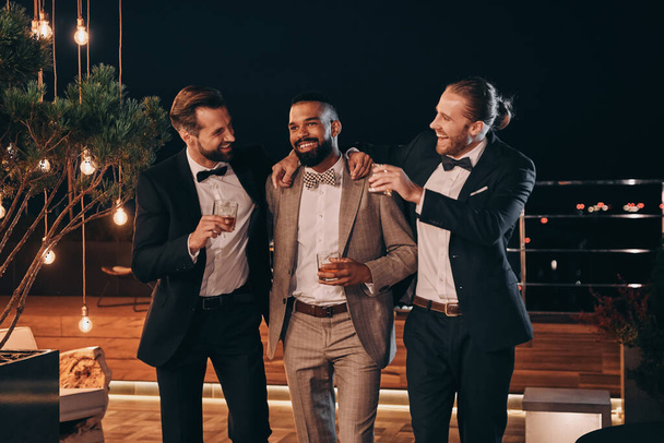 Three handsome men in suits drinking whiskey and communicating while spending time on party - Foto, Imagem
