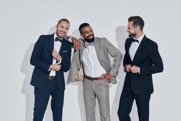 Three handsome young men in suits and bowties looking at camera and smiling while standing against gray background - Foto, Imagem