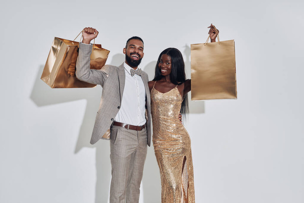 Beautiful African couple carrying gold colored shopping bags and smiling while standing against gray background - Foto, Imagem