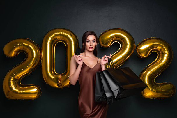 Woman in elegant dress hold credit bank card and shopping bags on black background, numbers air balloons studio. - Fotoğraf, Görsel