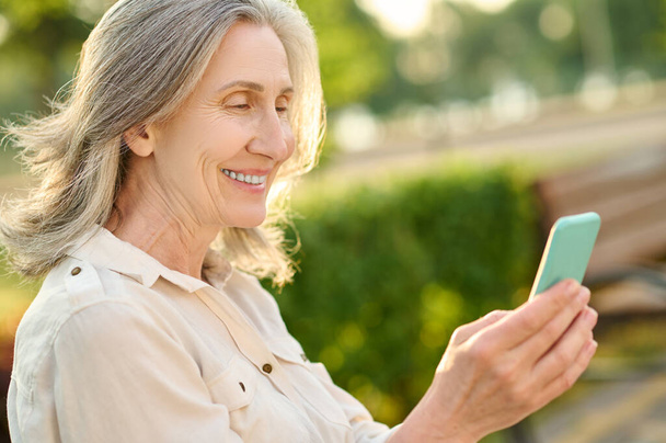 Smiling adult woman with smartphone in park - Φωτογραφία, εικόνα