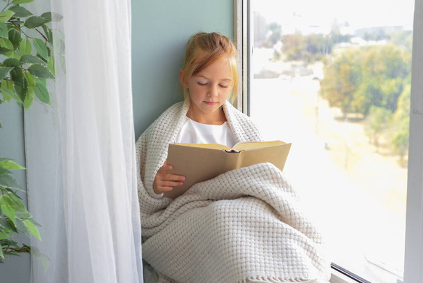 schattig meisje schoolmeisje het lezen van een boek door het raam thuis - Foto, afbeelding
