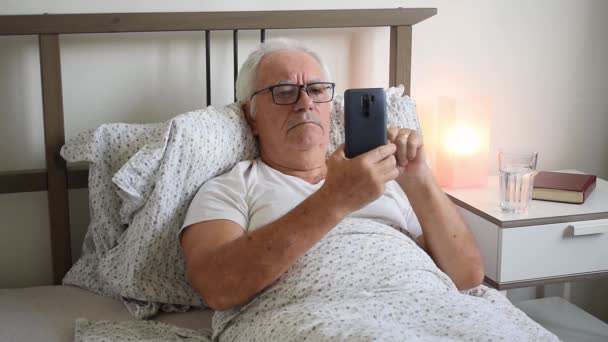ηλικιωμένος ηλικιωμένος άνδρας άνδρας στο κρεβάτι στο σπίτι επίπεδη, κουρασμένος άρρωστος μόνος συνταξιούχος και τον ιό ανάπαυσης φροντίδα δυστυχισμένος λυπημένος, κρατήστε smartphone - Πλάνα, βίντεο