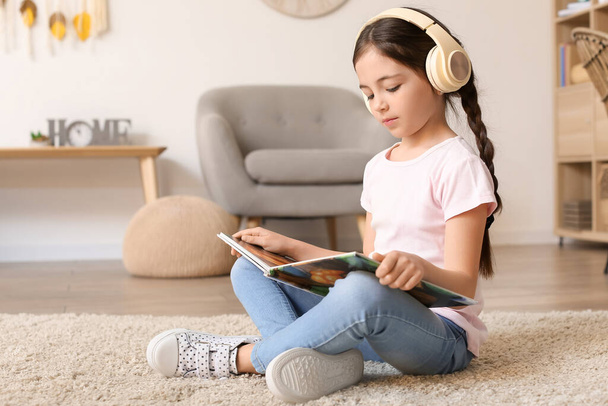Маленька дівчинка з навушниками читає книгу вдома
 - Фото, зображення