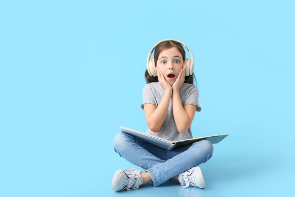 Surprised little girl with headphones and book on color background - Foto, Imagen