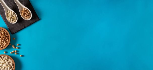 Ρεβίθια και φασόλια με κουτάλια πορσελάνης και επιφάνεια σχιστόλιθου σε μπλε φόντο. Επίπεδη. Αντιγραφή χώρου. - Φωτογραφία, εικόνα