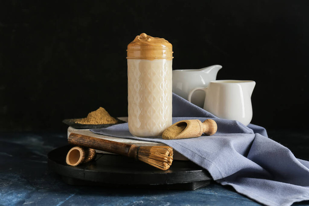 Ποτήρι νόστιμο dalgona hojicha latte, σέσουλα με σκόνη και chasen σε σκούρο φόντο - Φωτογραφία, εικόνα