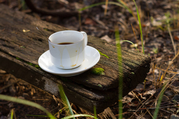 A white cup with a spoon on a white saucer stands on an old wooden board against a background of green grass. - Foto, immagini