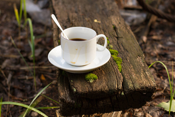 A white cup with a spoon on a white saucer stands on an old wooden board against a background of green grass. - Φωτογραφία, εικόνα