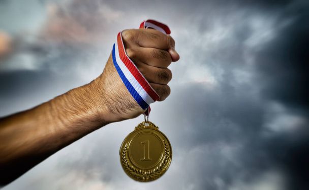 Hand holding gold medal against cloudy twilight sky background, in hand. First place award on cloudy sky background, Sport champion winner athlete and victory concept - Photo, Image