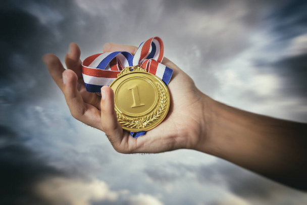 Medal for first place in the athlete palm against a dramatic sky. Winner gold award. Sport champion victory and success concept - Photo, Image
