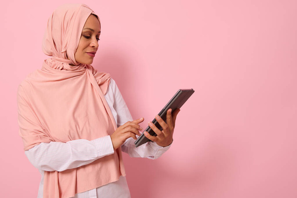 Beautiful Arab Muslim woman with covered head in pink hijab posing against a colored background with a digital tablet in hands, copy space for text. Advertising isolated portrait of a successful business lady using dogital electronic gadget - Fotoğraf, Görsel