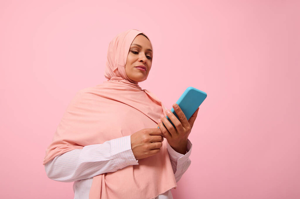 Mature Muslim woman of Middle Eastern ethnicity in pink hijab with smartphone in blue cover on her hands, isolated on pink pastel background with copy space for promotion - Fotó, kép