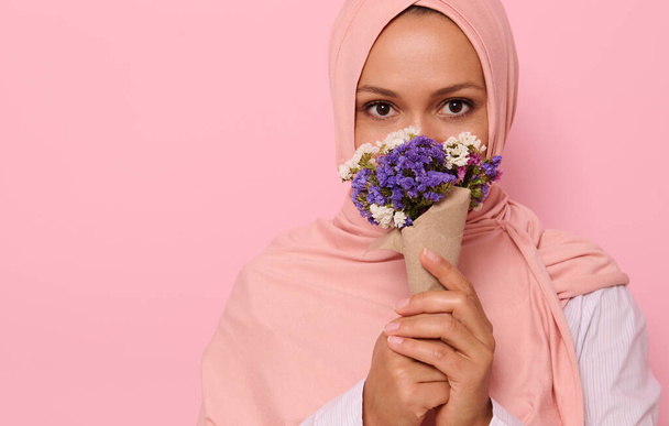 Close-up headshot portrait of young charming Arab Muslim woman in pink hijab with beautiful dark eyes, attractive gaze, looking at camera, covers half of face and mouth with a craft bouquet of flowers - Photo, Image