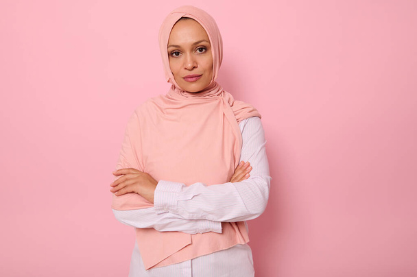 Waist-length portrait of beautiful Arabic Muslim woman in pink hijab posing looking to camera with attractive gaze, confident look and crossed arms, on pink background with copy space - Photo, Image