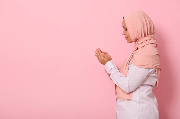 Retrato lateral de una hermosa y serena mujer árabe musulmana en hiyab rosa y atuendo estricto con palmas dobladas juntas a nivel de la cara realizando namaz, aisladas sobre fondo de color con espacio para texto - Foto, Imagen