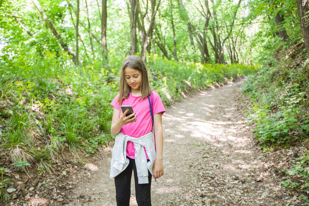 Μικρό κορίτσι πεζοπόρος χρησιμοποιώντας το τηλέφωνο για πλοήγηση στον αγροτικό δρόμο στο δάσος. - Φωτογραφία, εικόνα