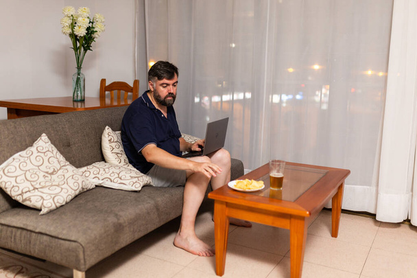 uomo barbuto sul divano utilizza computer portatile in sala da pranzo durante la notte - Foto, immagini